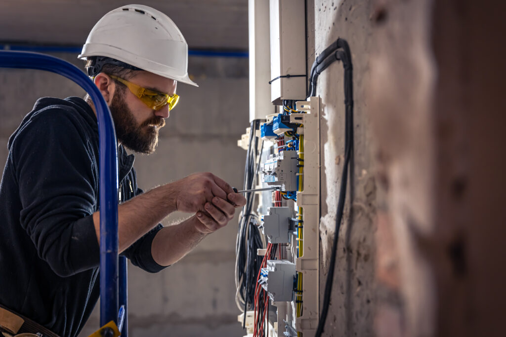 A male electrician works in a switchboard with an electrical connecting cable, connects the equipment with tools.